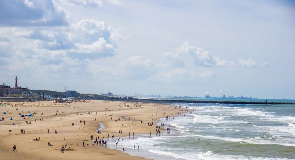 Panoramic view of the bustling Scheveningen Beach in The Hague with people enjoying the vast sandy shore and the dynamic waves, a lighthouse and pier in the distance, under a sky dotted with fluffy clouds.