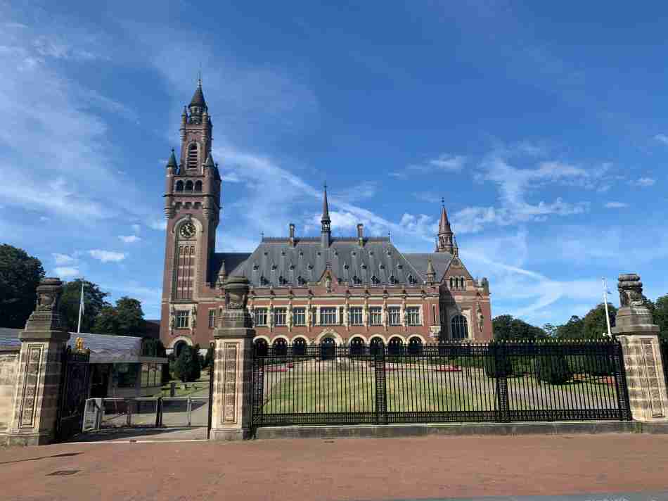 The iconic Peace Palace in The Hague, displaying its grandeur with a high clock tower and intricate architecture, surrounded by an ornate fence under a clear blue sky, symbolizing international law and order.