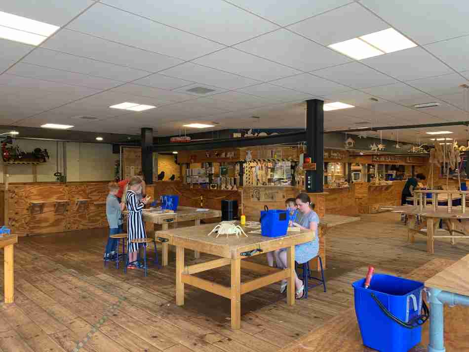 Children engaged in creative activities at the Hof van Saksen Academy workshop room, with wooden work tables and various craft materials, in a spacious and well-lit interior.