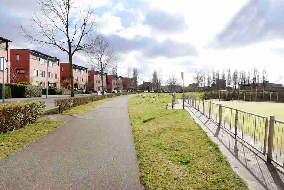 A winding pathway in Leidschenveen, a new neighborhood in The Hague, with a row of modern terraced houses on one side and a fenced sports field on the other, under a sky with dramatic cloud patterns.