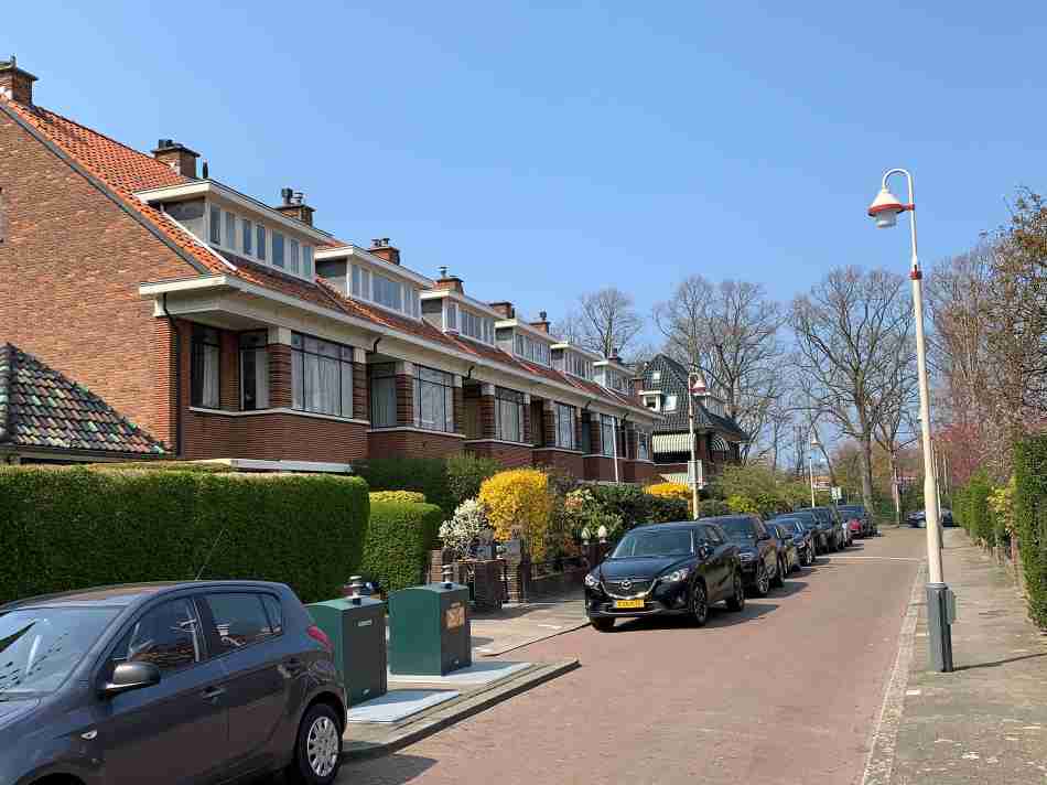 A sunny residential street in Benoordenhout, an upscale neighborhood in The Hague, with elegant semi-detached houses adorned with large windows and red tile roofs, neatly parked cars, and lush greenery enhancing the curb appeal.