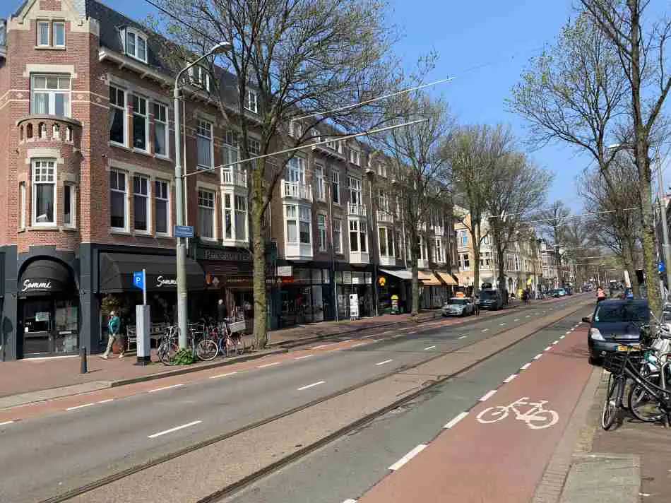 A vibrant street view in the Statenkwartier, one of the best neighborhoods in The Hague, showcasing a bike lane in the foreground, traditional Dutch architecture, and a variety of shops under a clear blue sky