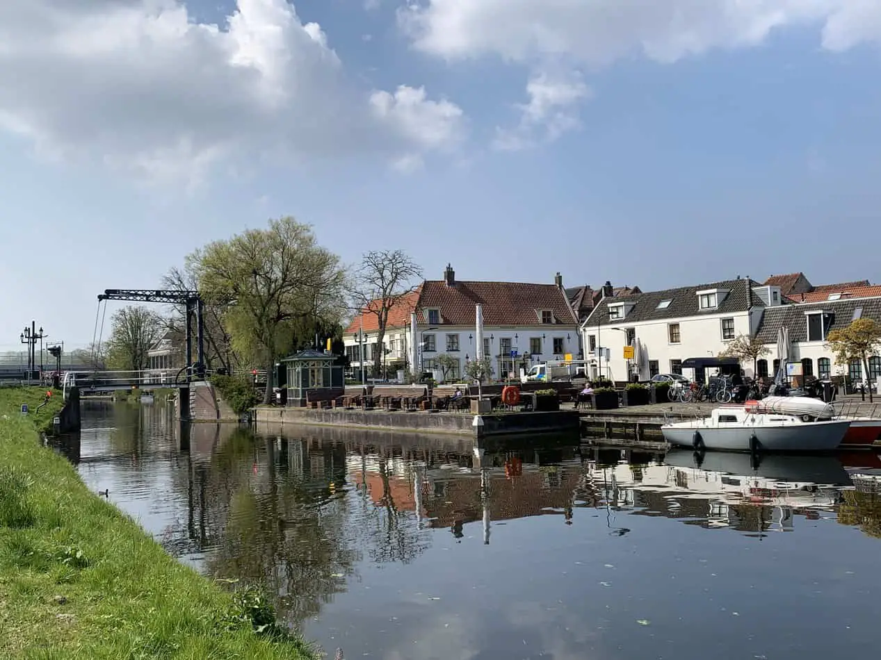 Idyllic canal view in Voorburg, a charming village adjacent to The Hague, featuring a traditional Dutch drawbridge, white moored boats, and quaint houses with outdoor seating, reflecting a serene lifestyle under a partly cloudy sky.