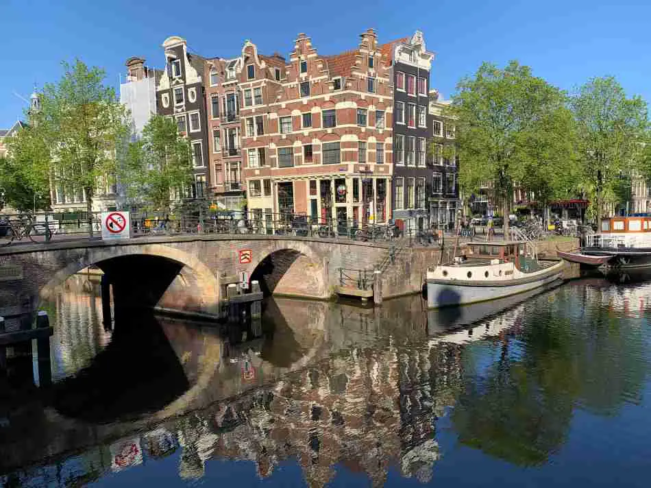 Historic corner of Prinsengracht and Brouwersgracht in Amsterdam, featuring ornate canal houses with gabled facades and the reflection of the buildings in the tranquil canal water, with a bridge and moored boats under a clear sky.