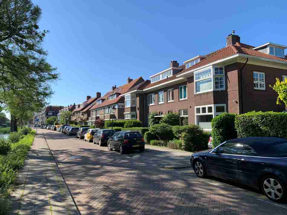 Clear blue skies over Hilligersberg, one of the best neighborhoods in Rotterdam, with a view of a peaceful street lined with mature trees and a neat row of classic Dutch houses with large windows and red-tiled roofs, accompanied by cars parked along the sidewalk.