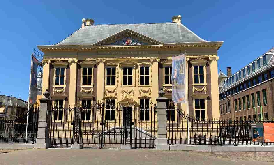 The Mauritshuis Museum in The Hague, a striking example of 17th-century Dutch classicism, with its honey-colored facade and ornate gilded details, secured behind a wrought-iron fence under a bright blue sky.