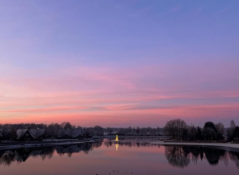 A peaceful winter sunset at Hof van Saksen, with soft pastel pink and blue hues streaking the sky and mirroring on the lake's surface, flanked by the silhouetted resort cottages and bare trees, creating a serene and frosty landscape
