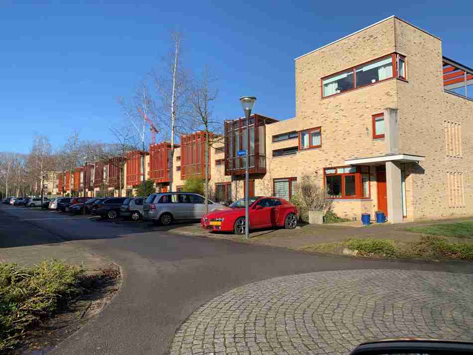 A sunny street view in Meerhoven, one of the best neighborhoods in Eindhoven for expats, displaying a row of contemporary town houses with distinctive orange accents and parked cars, reflecting the area's residential appeal