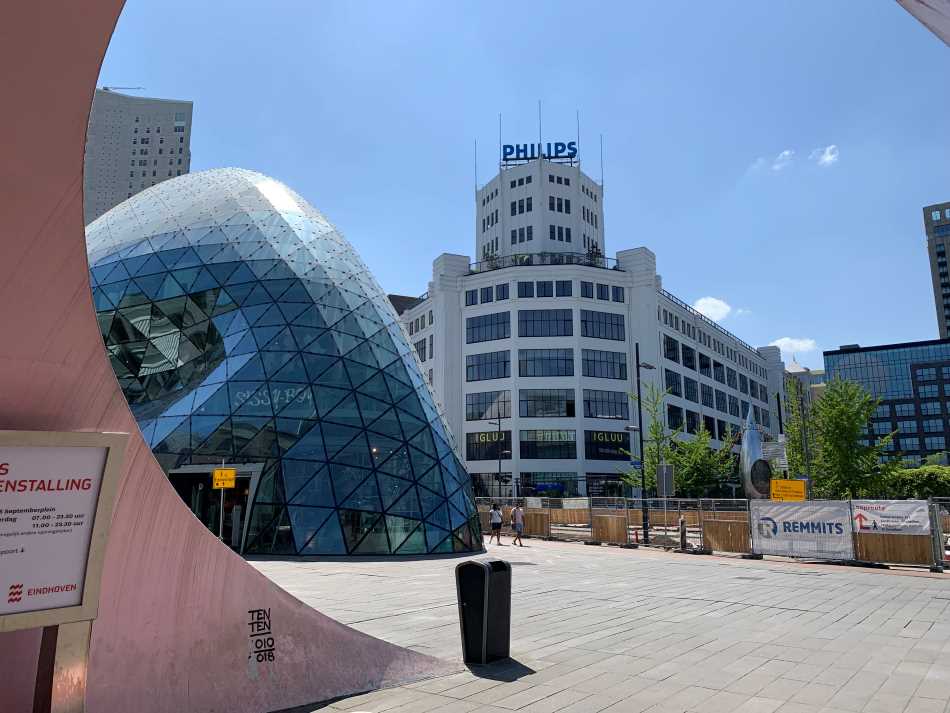 View of the juxtaposition of old and new architecture in the center of Eindhoven, featuring the iconic Philips building in the backdrop and a modern glass dome structure in the foreground, under a clear blue sky.