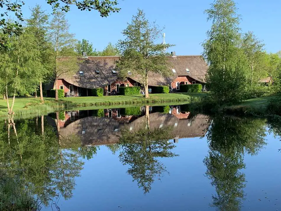 The serene Hof van Saksen cottages on a sunny day, with their reflections casting a mirror image on the still pond, surrounded by lush green trees, portraying a picture of tranquility and the resort's harmonious blend with nature